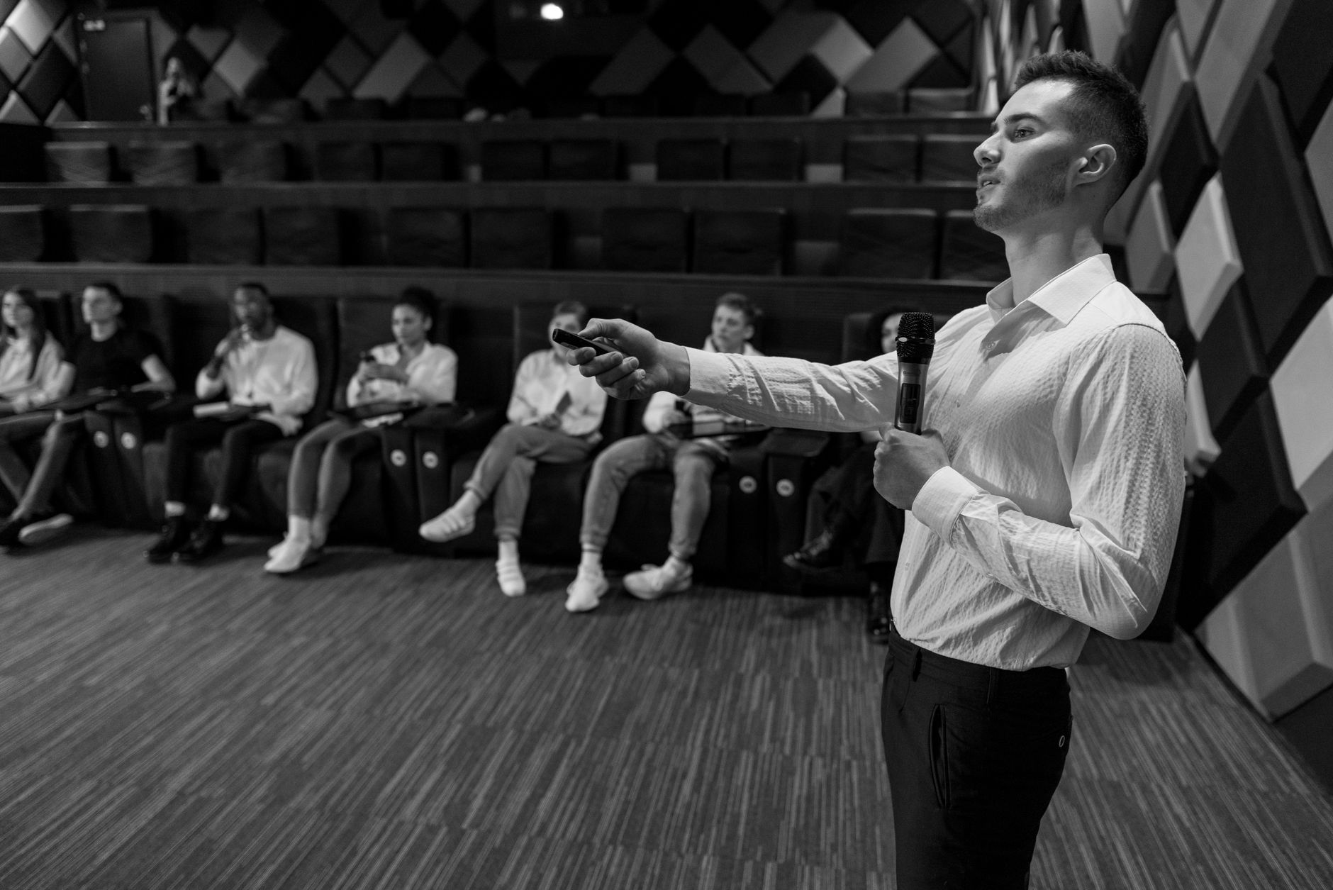 Monochromatic image of a speaker presenting to a small audience in a stylish auditorium.